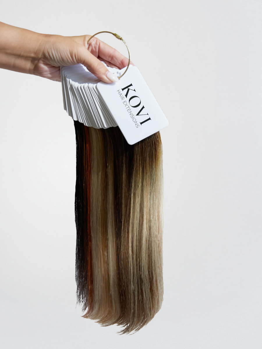 A professional KOVI Hair color ring held by hand, showcasing a full range of human hair extension swatches in various shades from light blonde to deep brunette. This essential tool helps stylists and clients select the perfect extension match for a seamless, natural blend.
